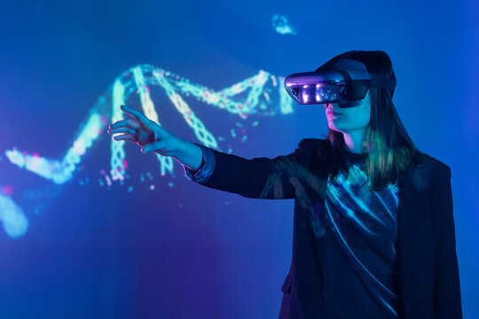 Side view of unrecognizable female with outstretched arm wearing VR headset while exploring virtual reality under blue neon light near wall with projector illumination