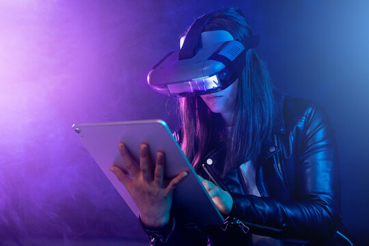 Concentrated anonymous female wearing modern VR headset standing near wall in dark room with contemporary tablet in hands under neon illumination