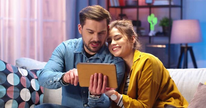 Portrait of joyful positive cute Caucasian couple watching at tablet screen and speaking surfing internet in good mood spending time together in evening at home sitting on sofa, family leisure concept