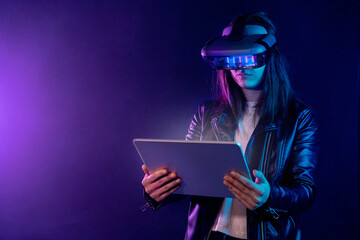 Concentrated anonymous female wearing modern VR headset standing near wall in dark room with contemporary tablet in hands under neon illumination