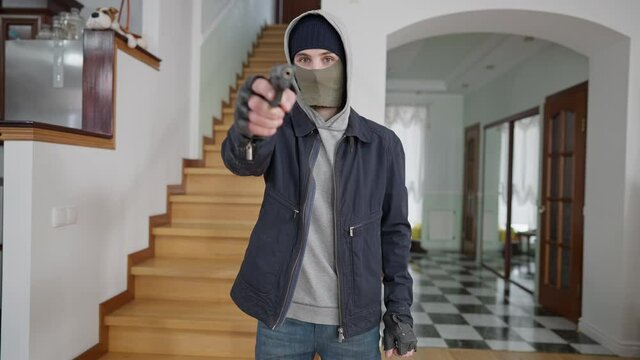 Middle shot portrait of young Caucasian masked man raising hand with gun looking at camera. Criminal robber threatening rich house owner with weapon. Burglar aiming at camera indoors.