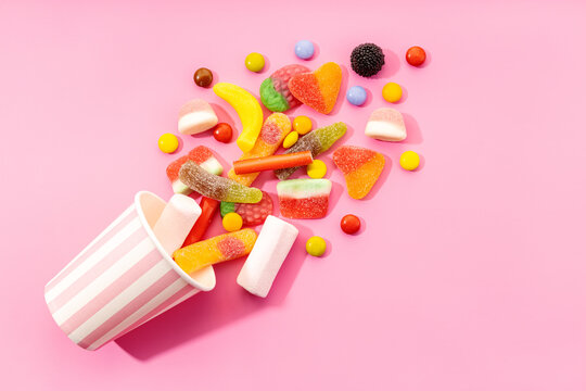 Top view of assorted multicolored sweet jelly candies and marshmallows scattered near disposable striped cup on pink background