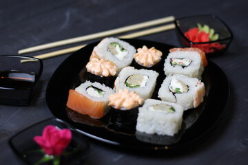 Pink rose in small bowl, marinated ginger and wasabi, soy sauce, rice rolls with salmon, crab, shrimp in plate and chopsticks on black background