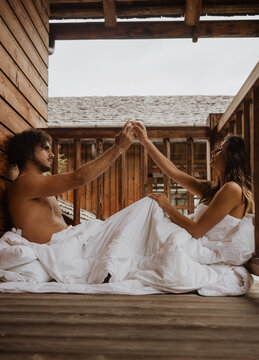 Side view loving young couple touching hands and looking at each other while sitting on terrace in soft white blanket