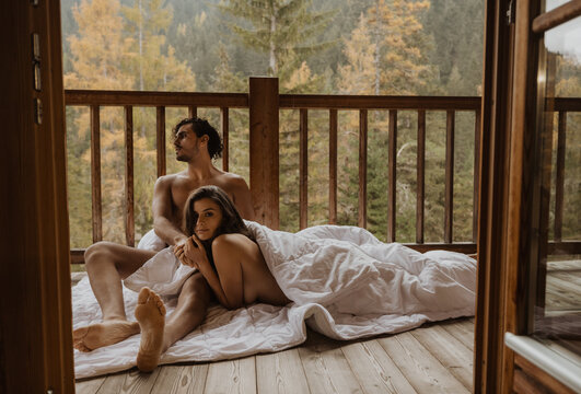 Romantic undressed couple lying on cozy blanket and cuddling on wooden cottage terrace against deciduous forest on autumn
