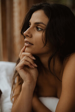 Attractive young brunette covering body with cozy blanket and touching face tenderly while looking away