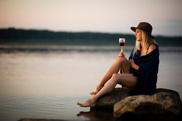 blonde girl in a hat with a glass of wine sitting on the riverbank, selective focus