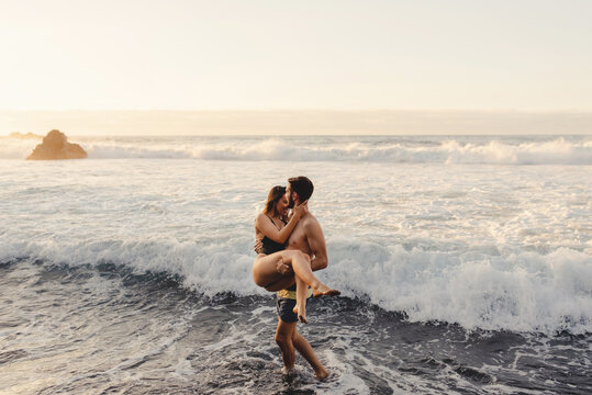 Full length of cheerful young man carrying girlfriend and walking out of waving sea water while spending sunset time together on beach of Tenerife island