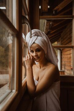 Young gentle female in towels touching face while looking at camera in wooden cabin