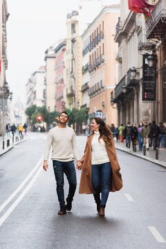 Full length content young couple in warm clothes holding hands and walking together on empty road in contemporary city on clear autumn day