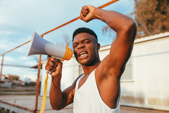 Young angry African American male in undershirt with speaker yelling with raised arm while looking at camera