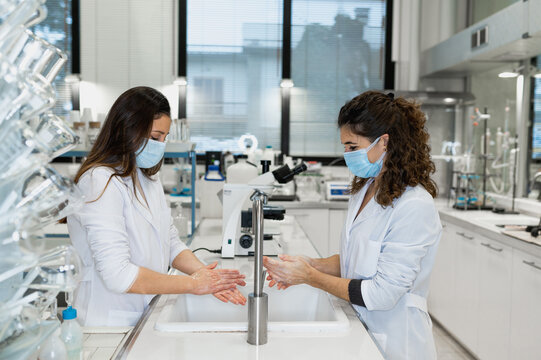 Side view of female scientists in white coats and protective masks washing hands before conducting chemical experiment in modern laboratory