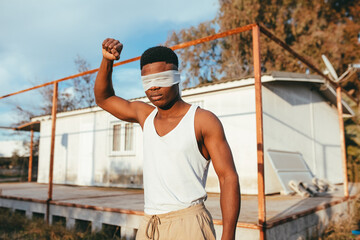 Anonymous masculine ethnic male protester in undershirt and blindfold standing with raised arm in countryside