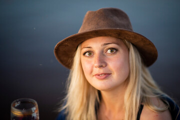 blonde girl in a hat with a glass of wine sitting on the riverbank, selective focus