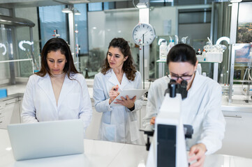 Group of professional female scientists using microscope and laptop while conducting research together in modern lab
