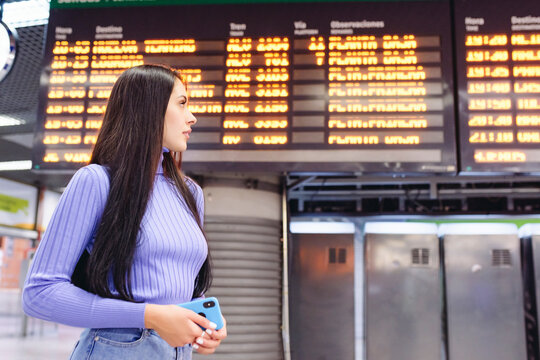 Side view of happy young ethnic female traveler in casual clothes standing in modern railway station near train schedule board holding smartphone