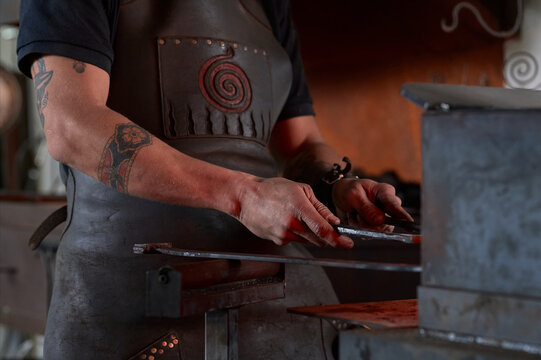 Side view of anonymous tattooed young male forger in apron heating metal in furnace while working in grungy smithy