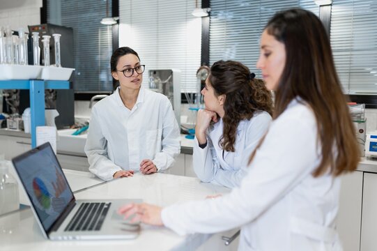 Young female pharmacy researchers gathering at table with laptop with diagrams and discussing results of research while working together in scientific lab