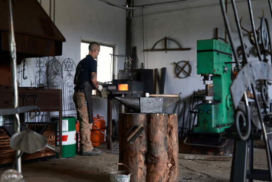 Back view of serious tattooed young male forger in apron heating metal in furnace while working in grungy smithy