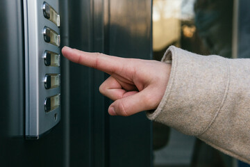 Crop anonymous female in warm coat pressing entry phone button while standing near building entrance door on clear autumn day