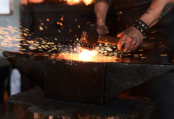 Side view of young brutal bearded craftsman in apron and goggles heating and striking iron on anvil with hammer during work in traditional smithy