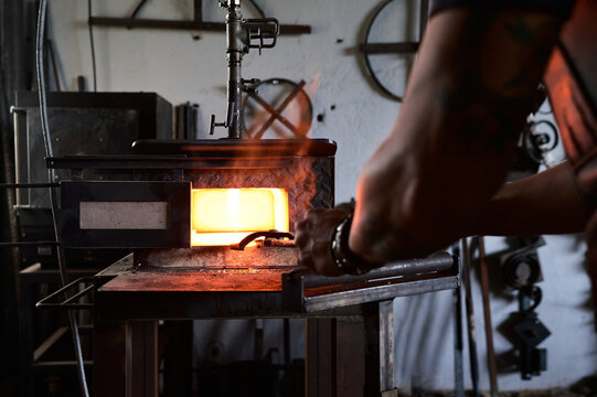 Back view of anonymous tattooed young male forger in apron heating metal in furnace while working in grungy smithy