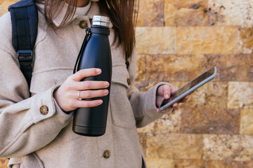 Crop anonymous female in warm coat with backpack holding reusable water bottle and browsing modern mobile phone against building wall on street