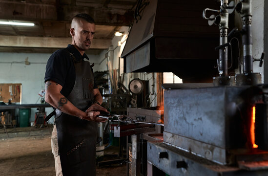 Side view of serious tattooed young male forger in apron heating metal in furnace while working in grungy smithy