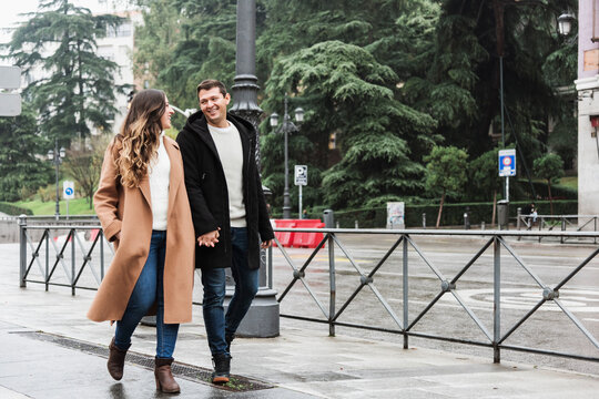 Full length cheerful young couple wearing warm clothes holding hands and looking at each other with smiles while walking together on asphalt sidewalk in modern city