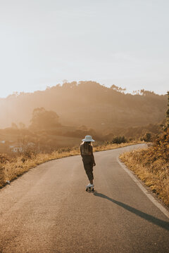Back view faceless sporty female in trendy wear riding cruiser board along empty asphalt road in summer countryside on sunny day