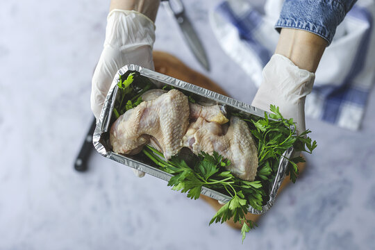 From above of faceless cook in gloves holding foil container with parsley and raw chicken thighs