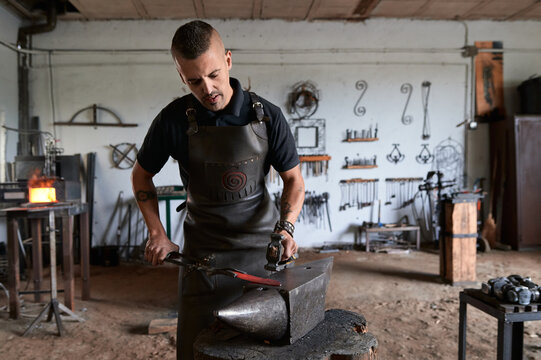 Front view of serious tattooed young male forger in apron heating metal in furnace while working in grungy smithy