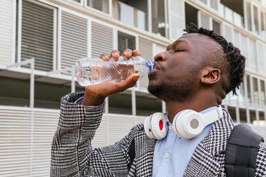 Young African American male manager with headphones and closed eyes drinking water from bottle against building in town