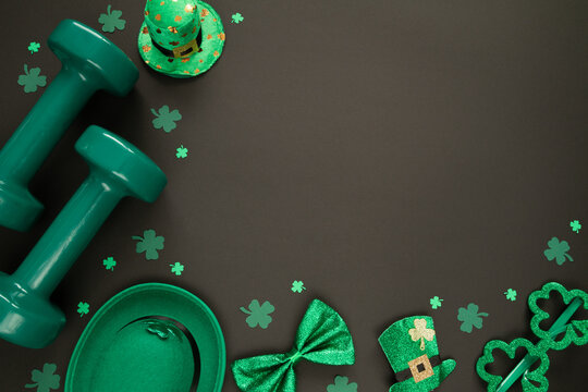 Two heavy green dumbbells, Irish hats with leaf clover, shamrock shaped glasses and a bow tie. Healthy fitness gym flat lay composition concept for St. Patrick's Day. Copyspace on black background.