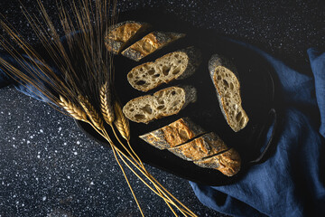 From above of appetizing crusty bread near wheat spikes and dark fabric on table