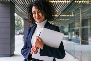 Modern successful well dressed ethnic female in elegant formal wear with laptop in hands smiling and looking at camera while standing near contemporary business building