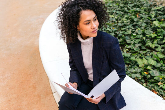 From above content young ethnic female entrepreneur in stylish formal suit studying documents in folder while sitting on city street looking away