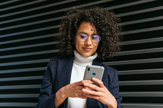 Smiling young ethnic curly haired female in stylish outfit browsing on mobile phone against gray wall