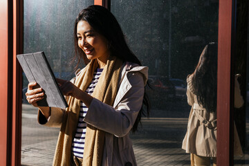 Cheerful Asian female standing against reflecting mirrored wall and browsing gadget on sidewalk in sunny day