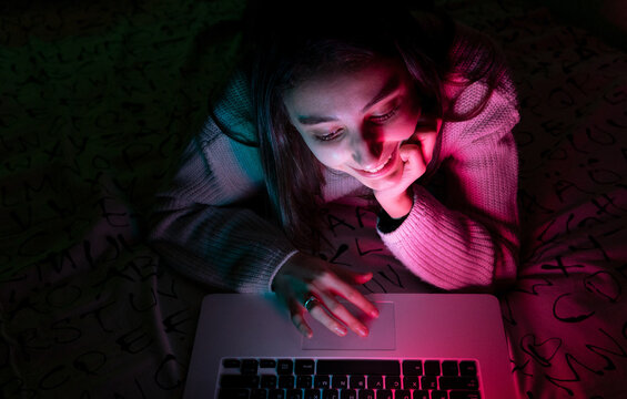 Smiling teenager in casual sweater lying on bedroom while browsing netbook in dark room