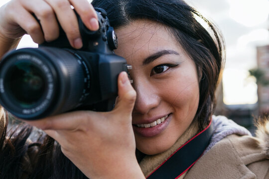 Ethnic young happy Asian female photographer shooting photo on professional photo camera on city street