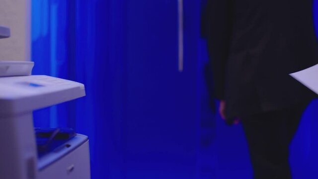 Man using  laser jet printer or scanner and taking an A4 paper document . Man in black suit opening  modern glass door and going out from office room with blue walls . Using copier in blue background