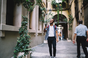 Smiling handsome dark skinned millennial hipster guy traveler, walking in historic center of city listening online audio guide in earphones connected to smartphone,positive male meloman enjoying music