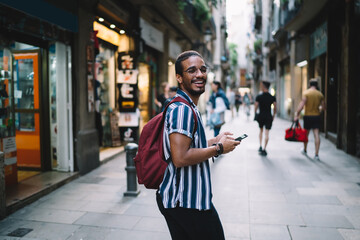 Happy african american male tourist laughing and walking on city street with smartphone on holiday vacations, positive dark skinned hipster guy looking at camera strolling with mobile phone on journey