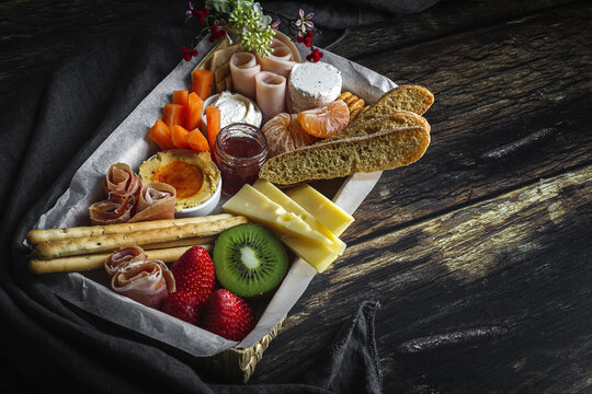 From above brunch box with assorted sliced meats various types of cheese and crispbreads arranged near ripe cup kiwi sweet strawberries and peeled mandarin near jam in glass jar on wooden table near napkin