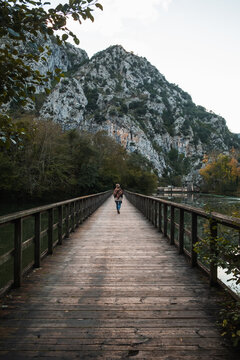 Distant tourist strolling on narrow wooden pathway above tranquil lake towards rough severe mountain ridge in Asturias