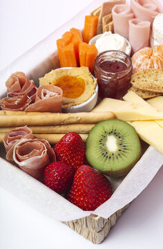 From above brunch box with assorted sliced meats various types of cheese and crispbreads arranged near ripe cup kiwi sweet strawberries and peeled mandarin near jam in glass jar on white background