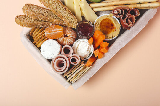 From above brunch box with assorted sliced meats various types of cheese and crispbreads arranged near ripe cup kiwi sweet strawberries and peeled mandarin near jam in glass jar on colorful pastel background
