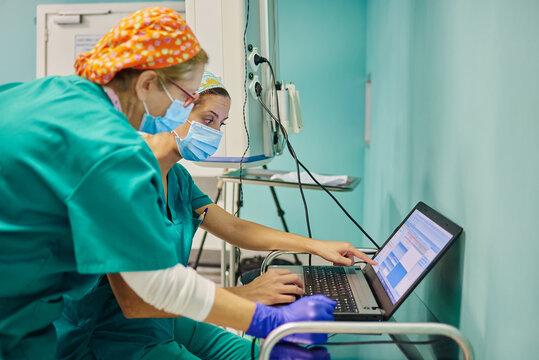 Side view of anonymous young females assistants in scrubs and masks using laptop during medical procedure in operating room