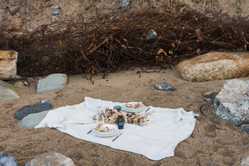 White blanket with food and decorations arranged for romantic picnic on sandy beach with stones on seashore